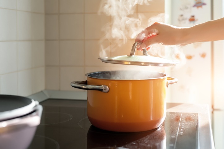 Female hand open lid of enamel steel cooking pan on electric hob with boiling water or soup and scenic vapor steam backlit by warm sunlight at kitchen. Kitchenware utensil and tool at home background